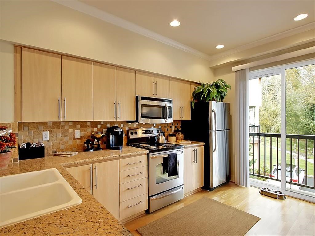 2-Bedroom Apartments in Kenmore, WA - Northshore Townhomes - Kitchen with Hardwood Flooring, Stainless Steel Appliances, Tan Wood-Tone Cabinetry, Slab Granite Countertops with Tile Backsplash, Decorative Plants, and Sliding Glass Door with Balcony Access.